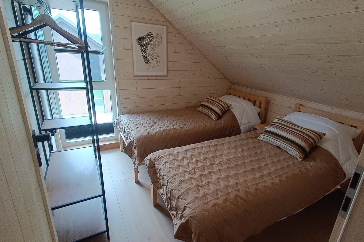 Jasna sypialnia z dwoma pojedynczymi łóżkami w domku nad jeziorem – drewniane ściany, skośny sufit, dekoracyjny obraz i regał z miejscem na ubrania.