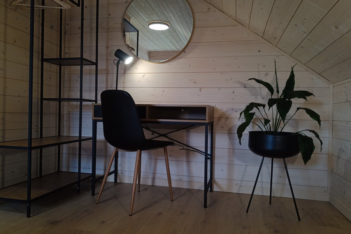 Przytulny kącik do pracy w domku wakacyjnym – minimalistyczne biurko z lampką, krzesło, okrągłe lustro i dekoracyjna roślina w czarnej donicy na tle drewnianych ścian.

