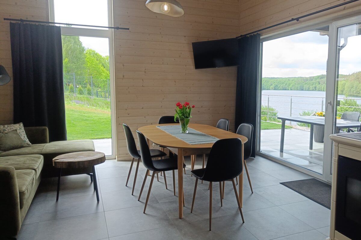 Nowoczesna jadalnia w domku letniskowym z widokiem na jezioro – przeszklone drzwi prowadzące na taras, stół z krzesłami, kwiaty i stylowy wystrój wnętrza z drewnianą boazerią.