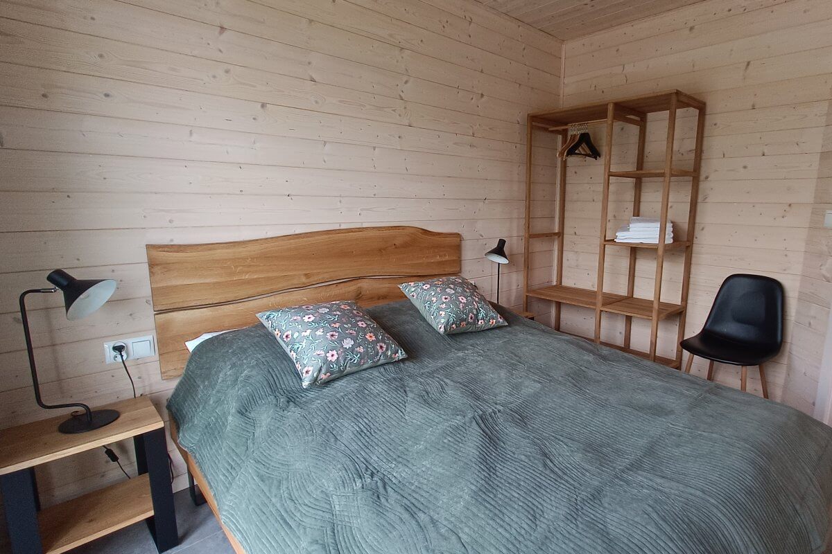 Drewniana sypialnia w domku nad jeziorem – podwójne łóżko z zieloną narzutą i kwiecistymi poduszkami, minimalistyczny regał na ubrania i stylowe oświetlenie.