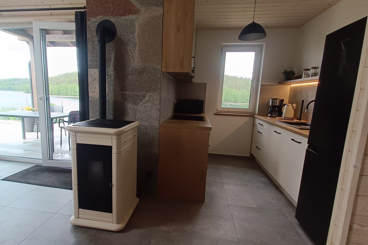 Przytulna kuchnia w domku wakacyjnym z widokiem na las – biała zabudowa, czarny sprzęt AGD, ekspres do kawy, okno z widokiem i nowoczesny kominek na pellet.