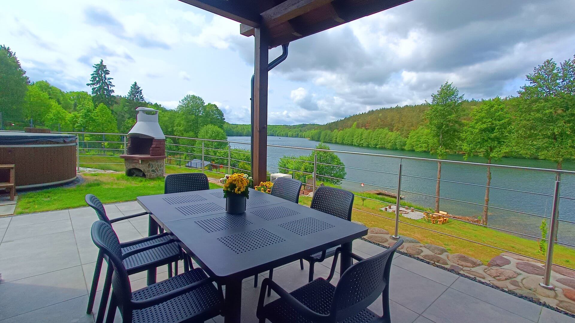 Zadaszony taras z widokiem na jezioro, czarny stół ogrodowy z krzesłami i grill w tle – Kaszubskie Tarasy