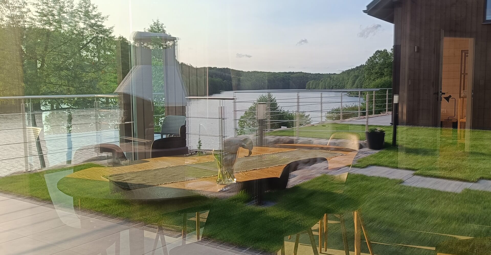 Widok przez szybę na taras nad jeziorem z designerskim stołem, krzesłami i kominkiem ogrodowym, z odbiciem wnętrza nowoczesnego domku letniskowego i panoramą zielonych wzgórz w tle.