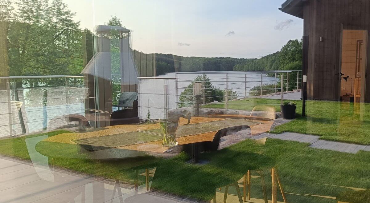 Widok przez szybę na taras nad jeziorem z designerskim stołem, krzesłami i kominkiem ogrodowym, z odbiciem wnętrza nowoczesnego domku letniskowego i panoramą zielonych wzgórz w tle.