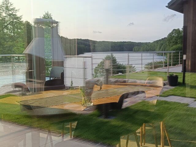 Widok przez szybę na taras nad jeziorem z designerskim stołem, krzesłami i kominkiem ogrodowym, z odbiciem wnętrza nowoczesnego domku letniskowego i panoramą zielonych wzgórz w tle.